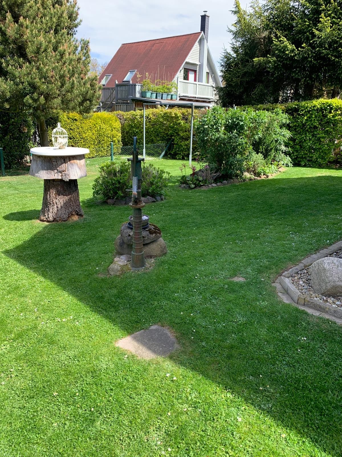 Garten mit Vogelhäuschen auf einem Baumstumpf und alter Wasserpumpe, dahinter das neue Klettergerüst aus Metall