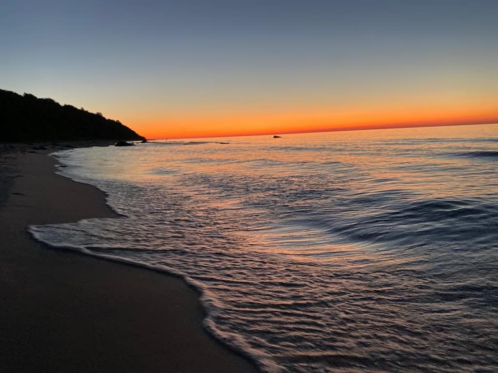Morgenrot über der Ostsee, flache Wellen am Sandstrand, links die bewaldete Küste
